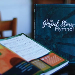 Photo of the Gospel Story Hymnal sitting on a table. One copy is closed so that you see the cover. Another copy is open showing an illustration of nature and the hymn "This is My Father's World."