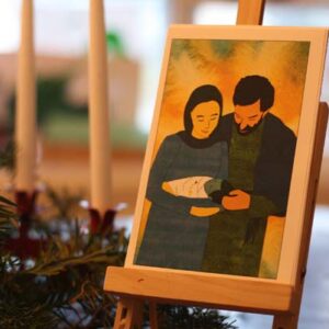 Photo of an advent card on an easel, next to an advent wreath. The card shows a color illustration of Mary and Joseph holding baby Jesus.