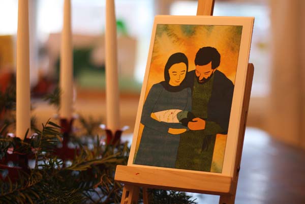 Photo of an advent card on an easel, next to an advent wreath. The card shows a color illustration of Mary and Joseph holding baby Jesus.