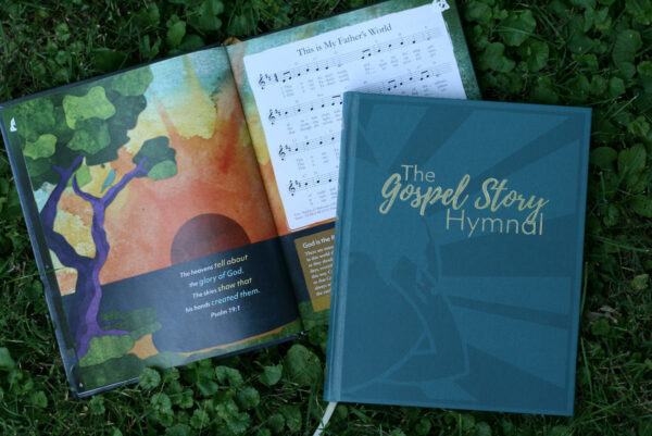 photo of the gospel story hymnal. One copy is open on the ground, showing a page with an illustration and a hymn. Another copy lays closed next to it, so you can see the front cover.